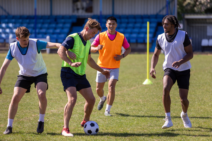 A medium close-up of a group of four young men playing a casual game of football They are all wearing different colored bibs over their athletic clothing, with two players wearing green and orange bibs and two others in white bibs.  One player in a green bib is actively trying to kick the soccer ball, The weather in Newcastle upon Tyne is sunny and warm, making it an ideal day for outdoor sports.

Videos are available similar to this scenario.